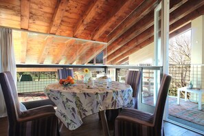 Eine Holzbalkendecke mit einer Glastür zum Balkon, ein runder Tisch mit einer Tischdecke mit Blumenmuster und gestreifte Stühle.