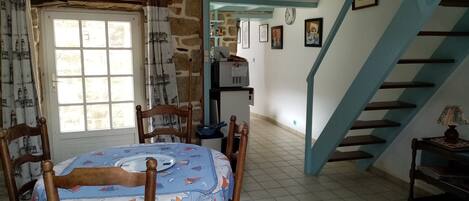 Un comedor con una mesa cubierta con mantel de temática náutica, sillas de madera, pared de piedra, escalera con detalles azules y una lámpara en el techo.