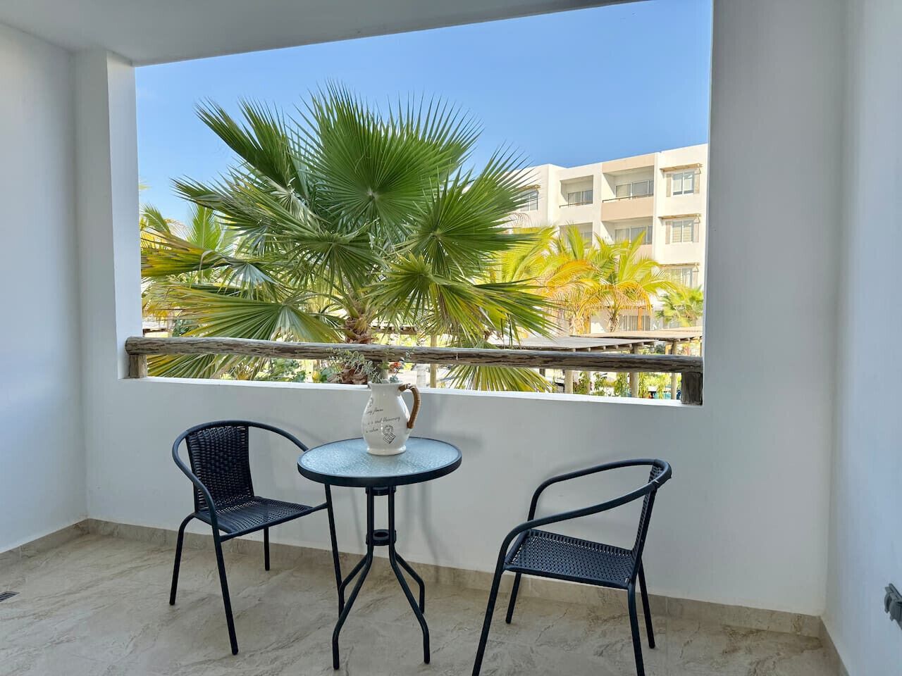 Un balcon avec deux chaises et une petite table, un pot de fleurs et une vue sur des immeubles et des palmiers.