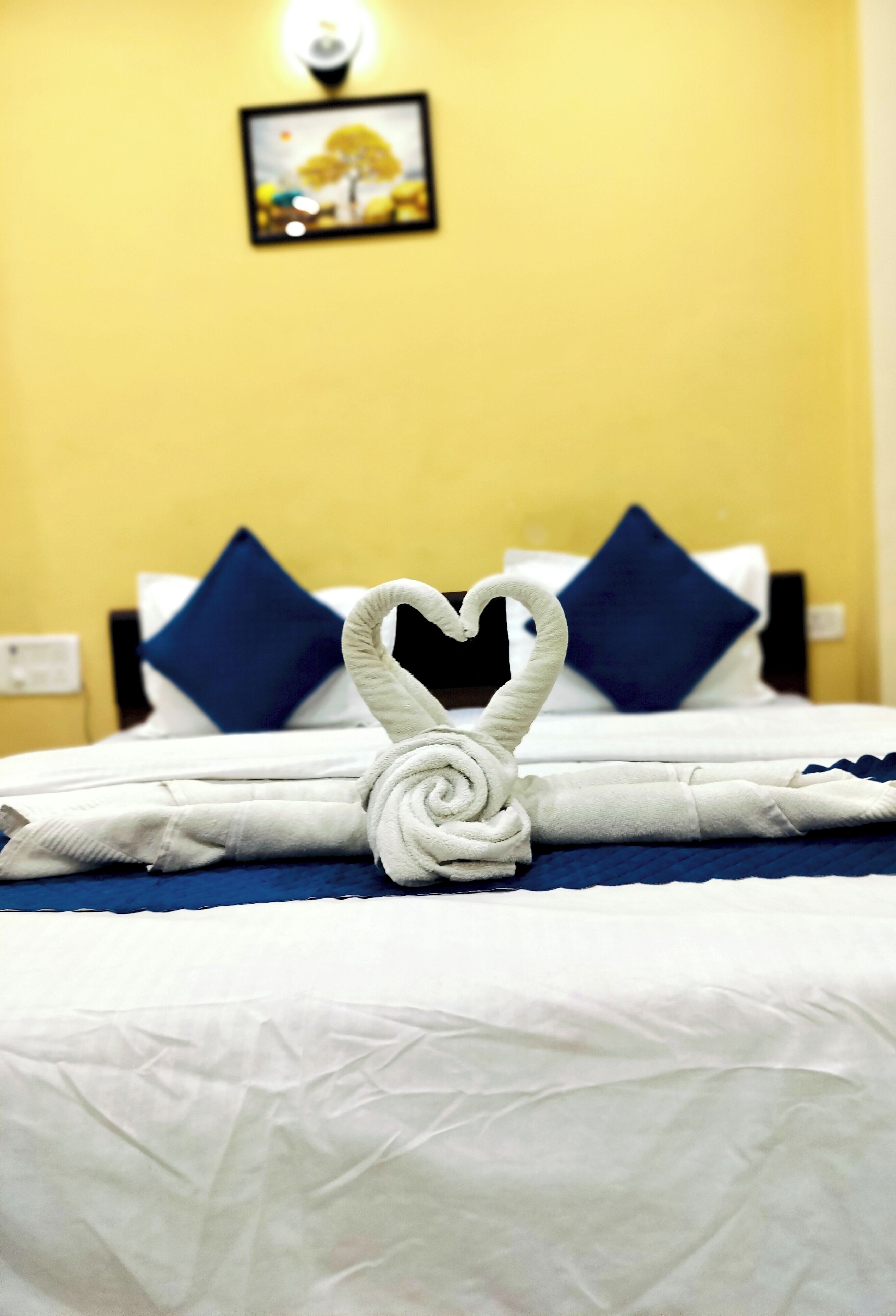A bed with a heart-shaped towel arrangement, a blue and white pillow, and a picture on the wall.