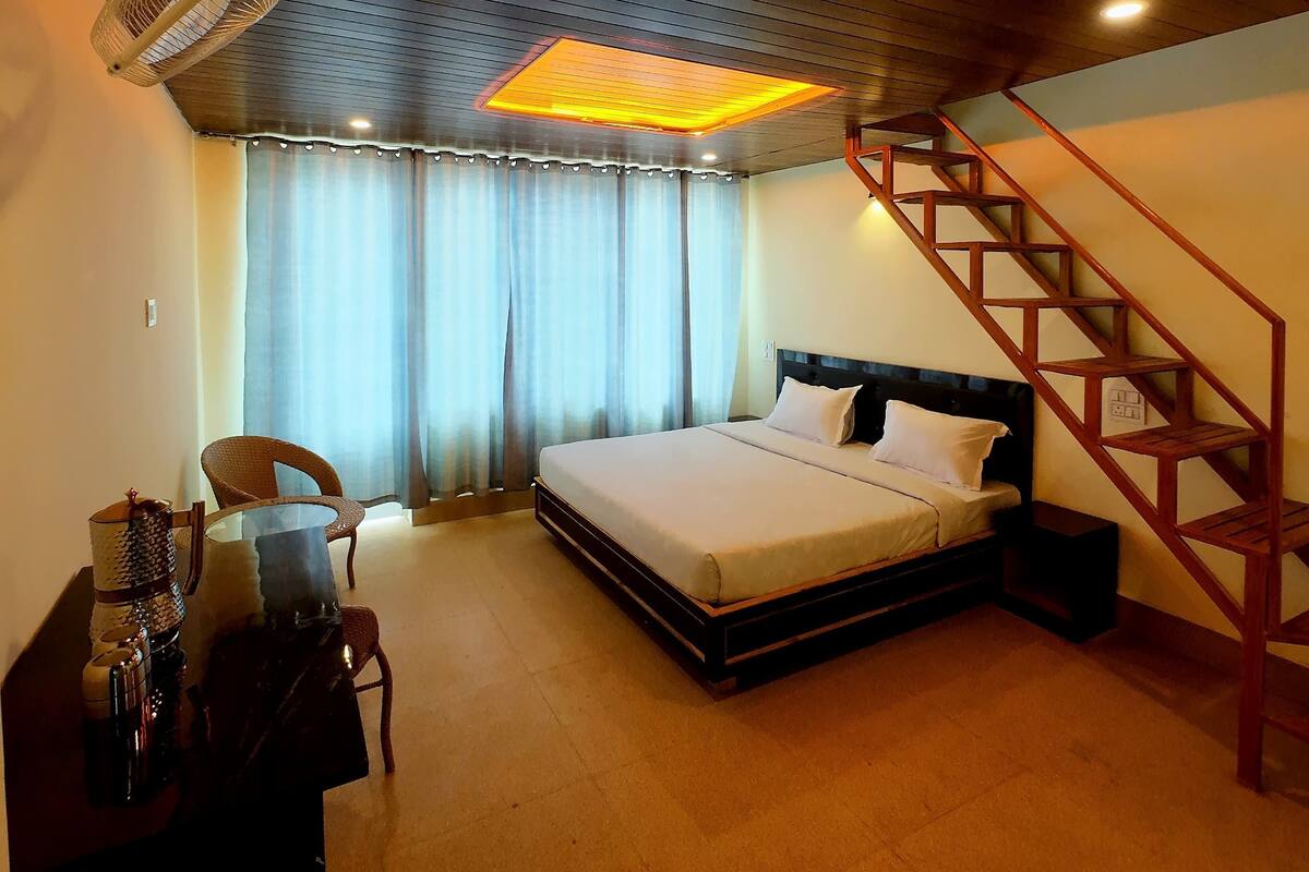 A bedroom with a bed, a chair, a desk, a standing fan, a staircase, and a ceiling light.