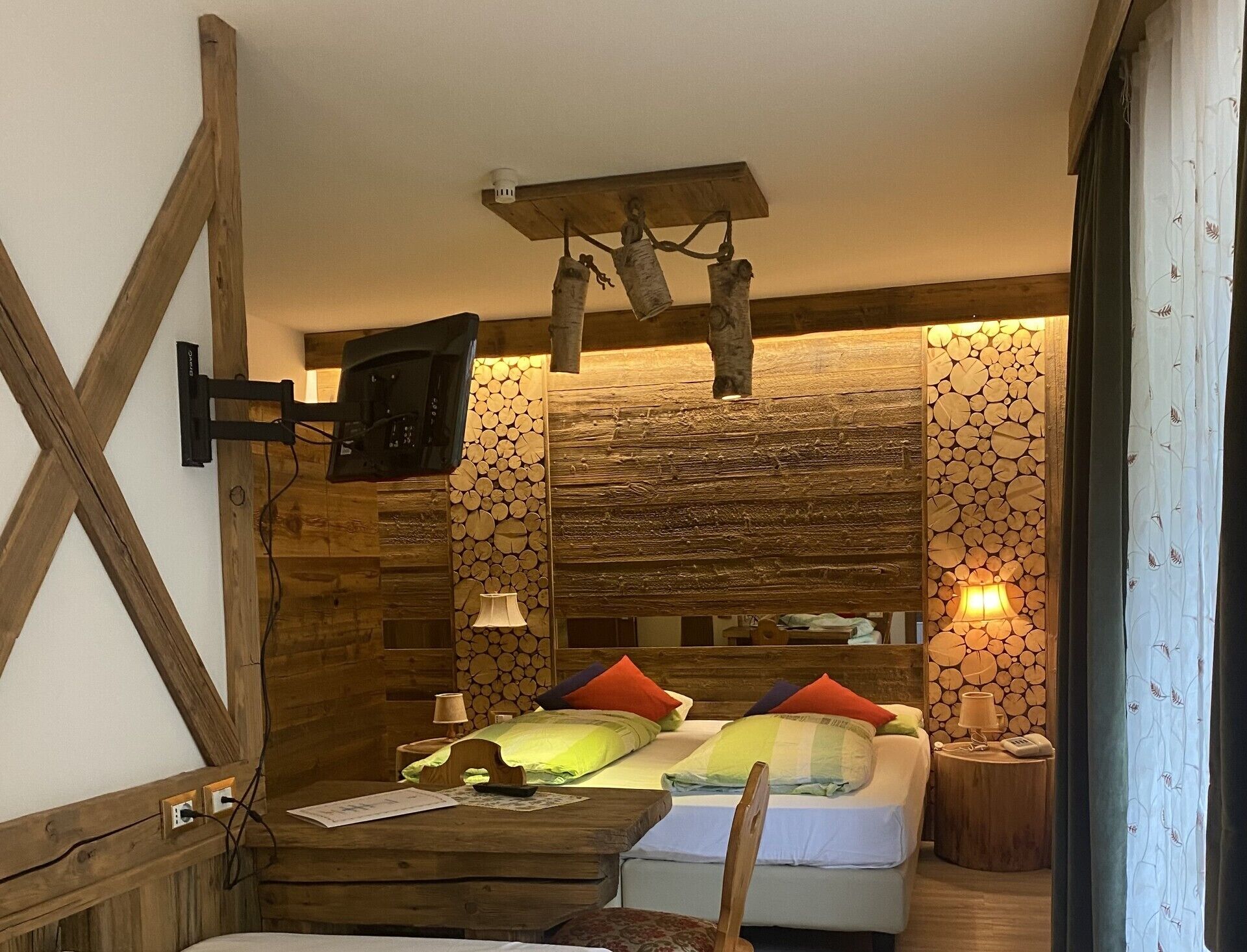 A cozy bedroom with a wooden headboard, a television mounted on the wall, a bedside table with a lamp, and a bed with colorful pillows.