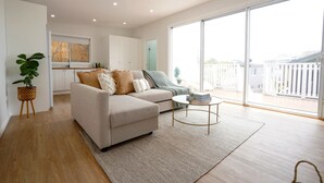A modern living room with a sofa, a coffee table, and a large sliding glass door leading to a balcony. - 5 mins walk to Beach - Modern Beachy Escape. Netflix, Full Kitchen, Comfy Beds! (Terrigal)