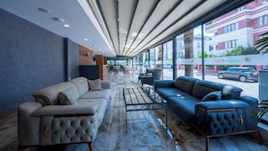 Un vestíbulo moderno con un sofá gris, un sofá azul y una mesa de centro. Hay ventanas amplias con vistas a la calle y edificios.