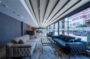 Un vestíbulo moderno con un sofá gris, un sofá azul y una mesa de centro. Hay ventanas amplias con vistas a la calle y edificios.