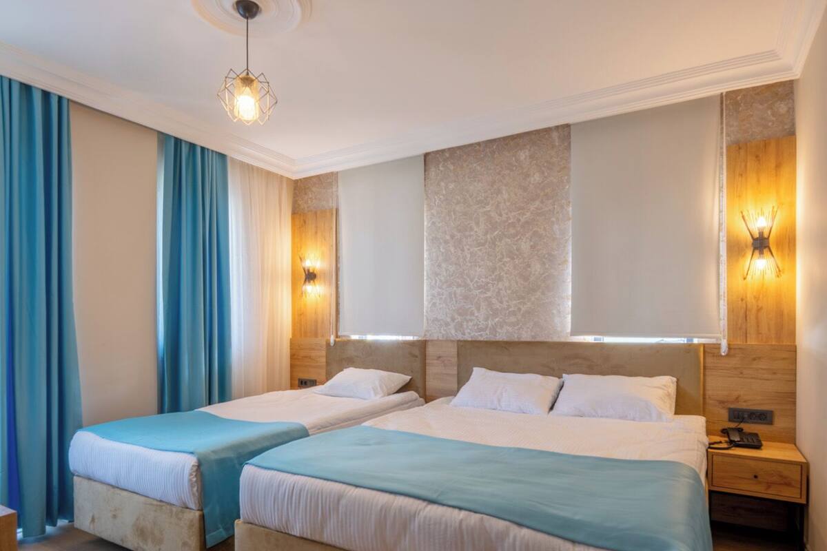 Habitación de hotel con dos camas, suelo de madera, un ventanal grande con cortinas y un aplique de pared.