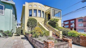 Un edificio amarillo con fachada curva, una escalera que conduce a la entrada y un pequeño jardín con plantas.