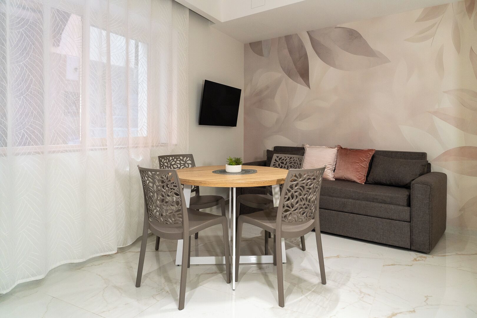 Un salon moderne avec une table ronde en bois, quatre chaises, un canapé gris et un téléviseur à écran plat fixé au mur.