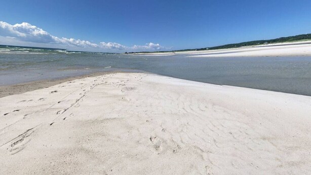 Een strand met voetafdrukken die naar het water leiden, een heldere lucht en een verre kustlijn.
