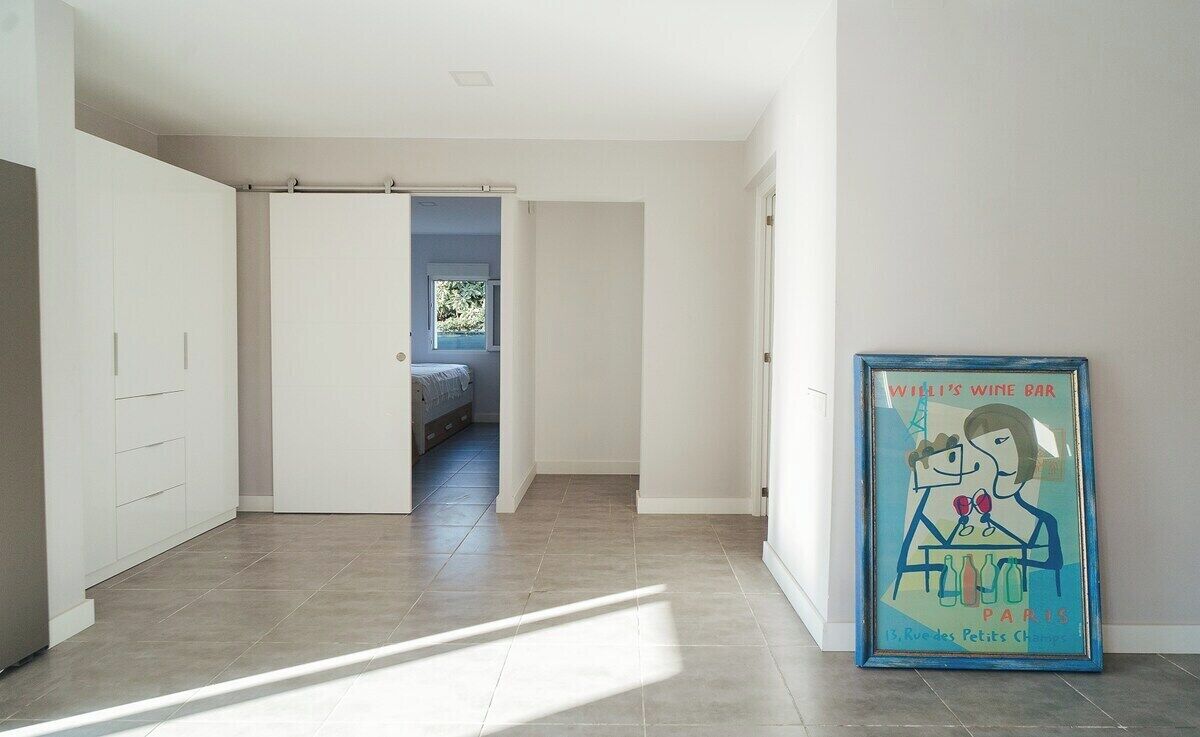 A hallway with a bed in the background, a framed picture on the wall, and a door leading to another room.
