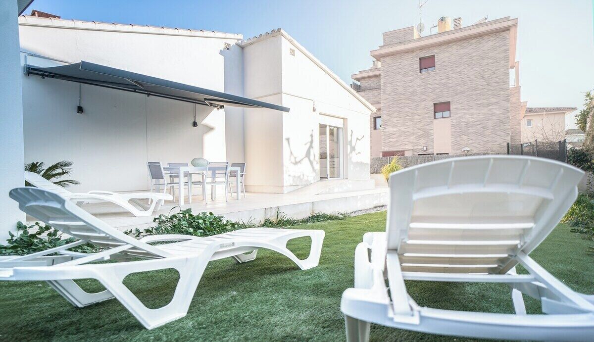 Un patio moderno al aire libre con sillones blancos y una mesa, rodeado de un césped cuidado y un edificio blanco con techo plano.