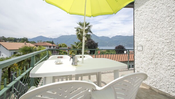 Un balcon avec une table et des chaises blanches, un parasol jaune, et une vue sur des montagnes et un lac.
