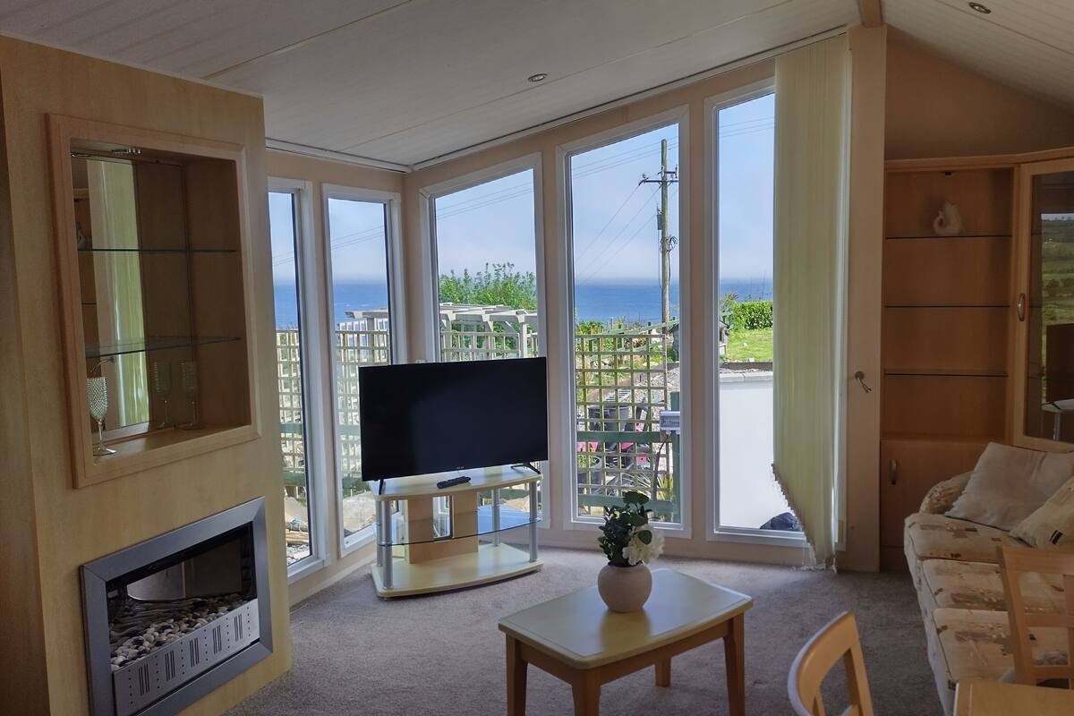 Ein großzügiger Wohnraum mit großem Fernseher, Kamin und Meerblick durch hohe Fenster.