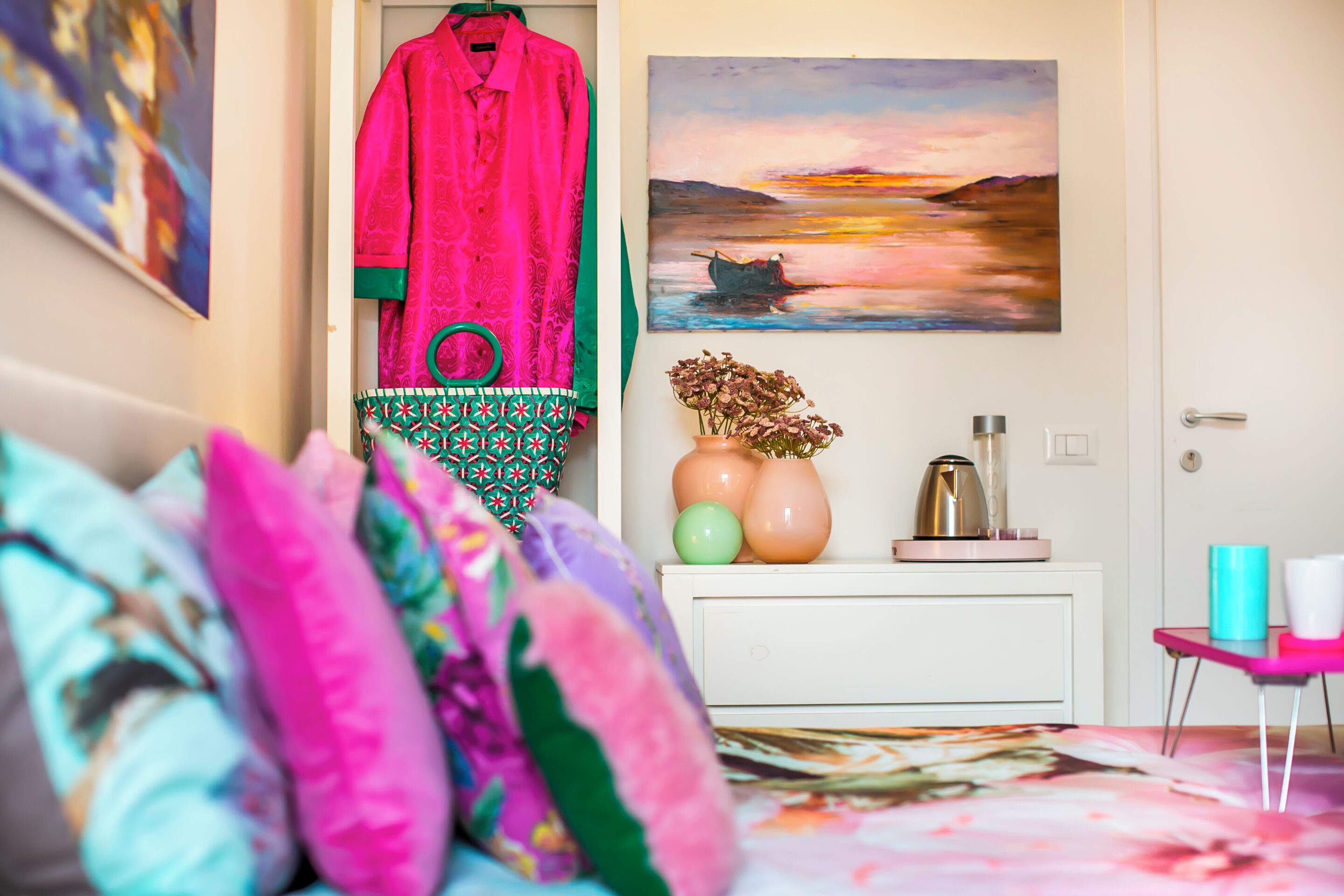 A bedroom with a bed, colorful pillows, a hanging garment, a painting, a vase with flowers, and a coffee maker.