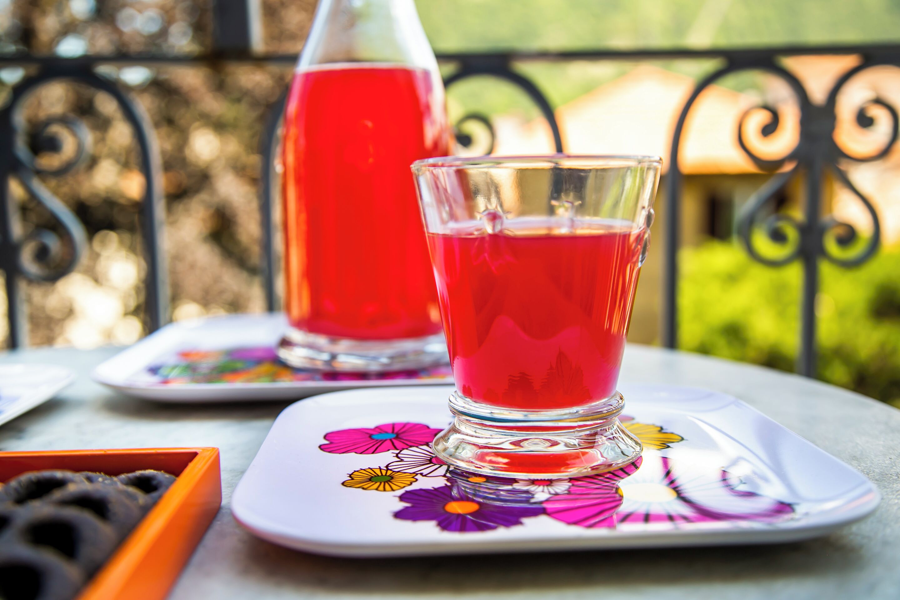 A glass of red beverage on a floral coaster, a bottle of red drink, and a tray with black objects on a table outdoors.