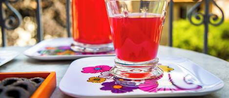 A glass of red beverage on a floral coaster, a bottle of red drink, and a tray with black objects on a table outdoors.