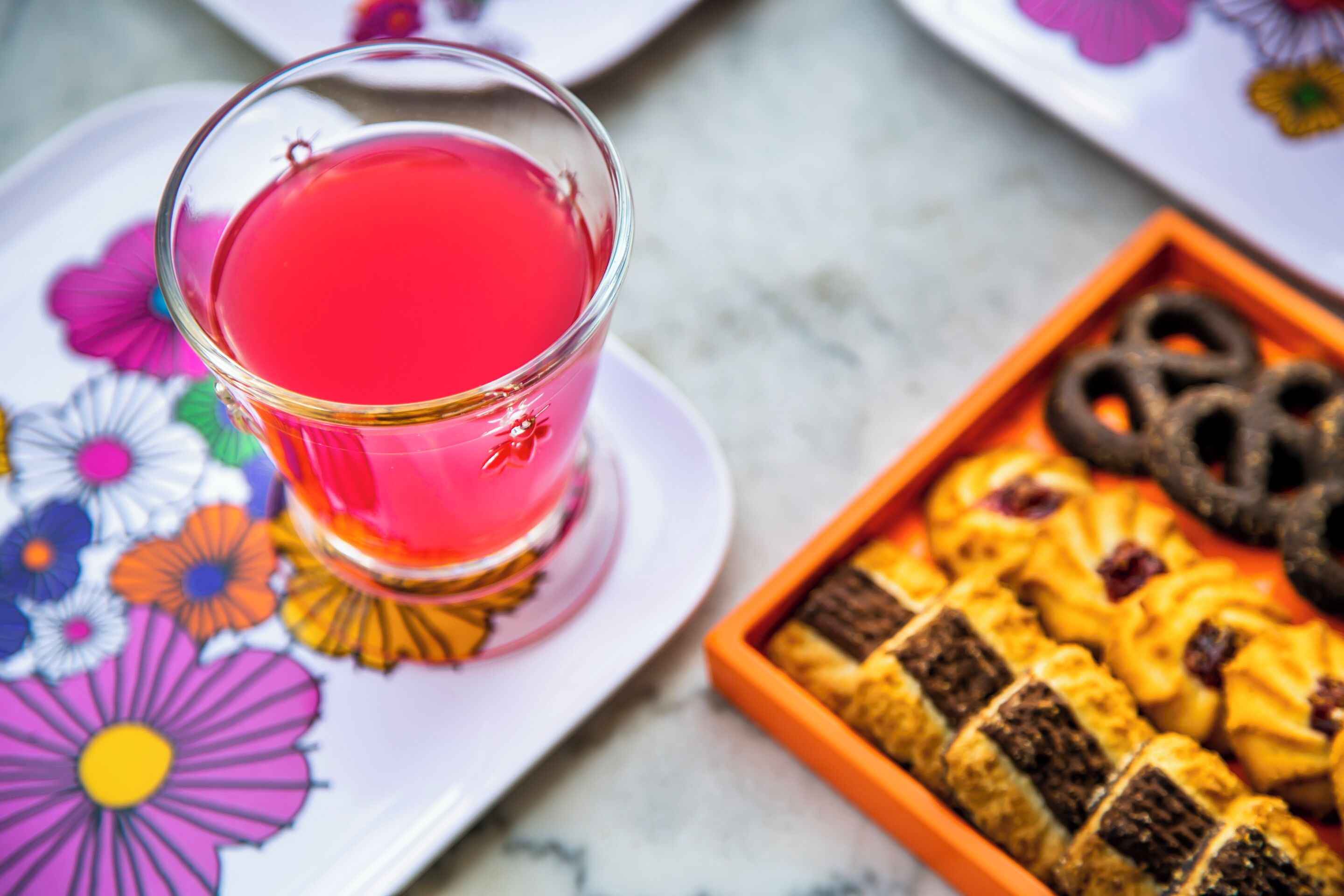 Un verre d’une boisson rouge sur une assiette à motifs floraux, un plateau de biscuits et une assiette décorative ornée de fleurs.