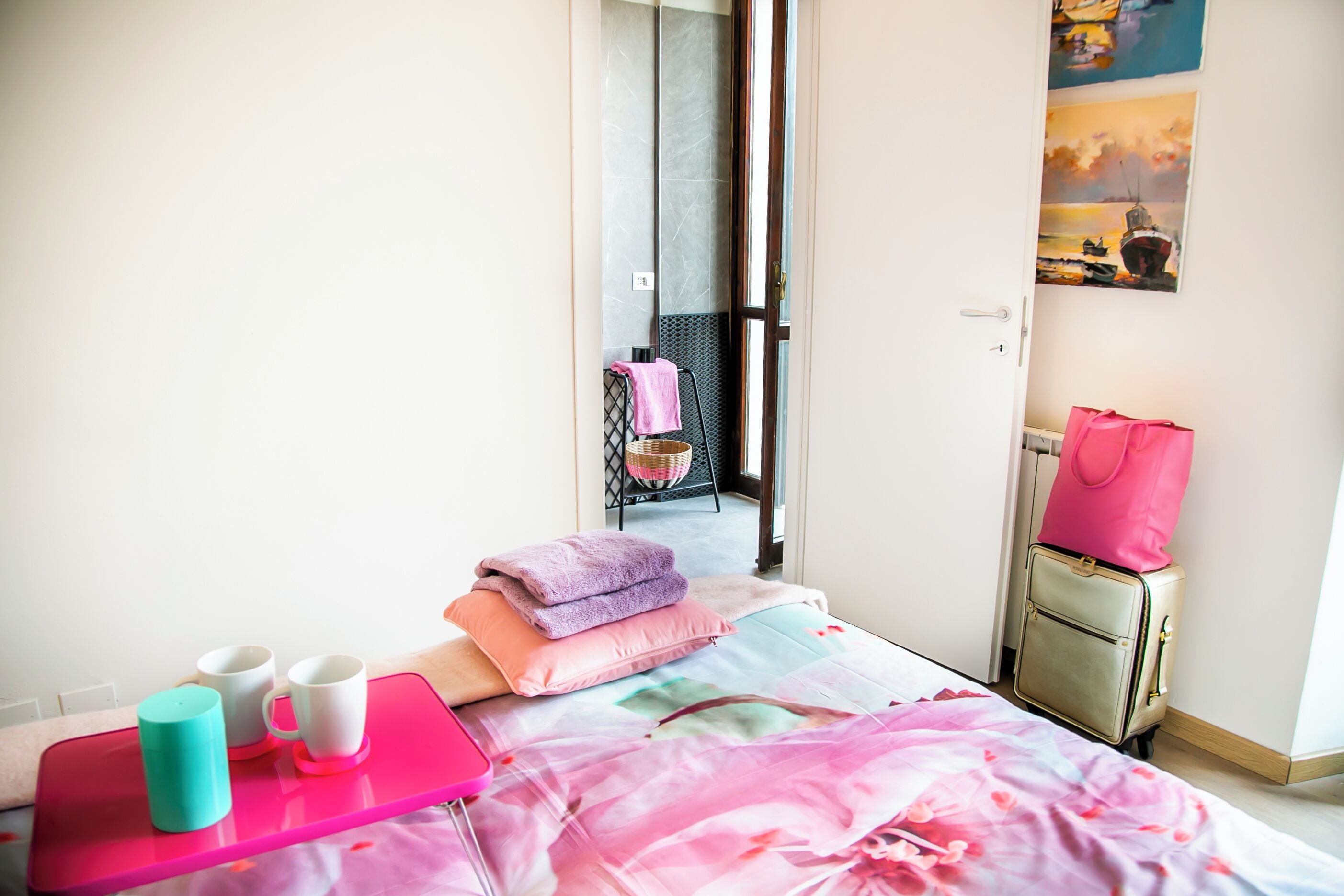 Una habitación de hotel con una cama, una bandeja rosa con tazas, una maleta y un cuadro en la pared.