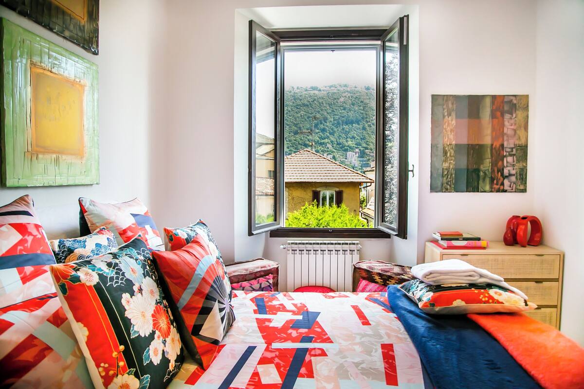 Ein Schlafzimmer mit großem Fenster, einem Bett mit bunter Bettw äsche, einem Heizkörper und einem kleinen Tisch mit Büchern und einem roten Gegenstand.