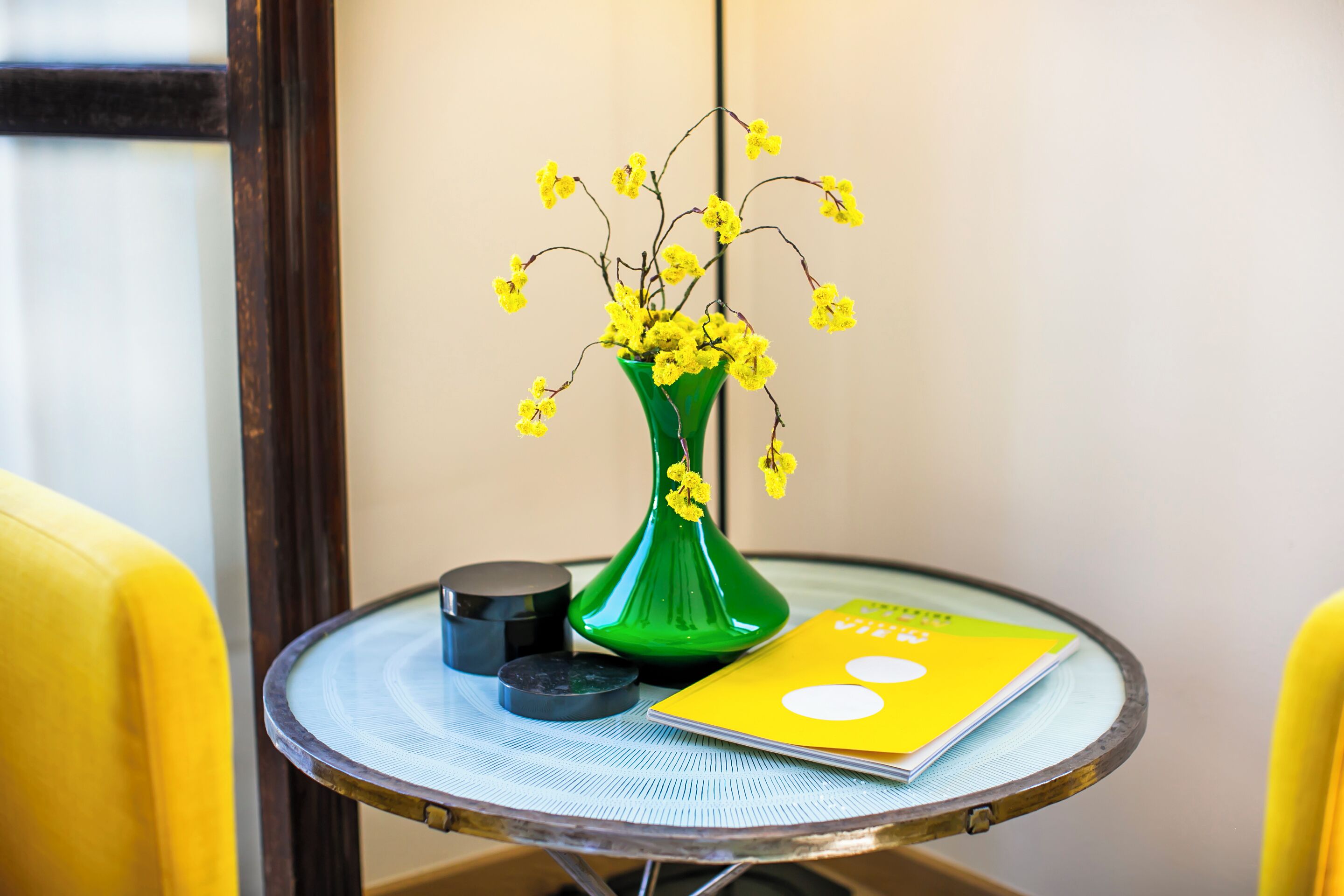 A round table with a green vase containing yellow flowers, a black object, and a yellow book.