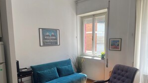 A room with a blue sofa, a rocking chair, a small table with a record player, and a window with a view of a building outside. - Central Boutique Apartment for2 (Roma)