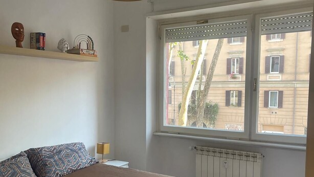 A bedroom with a bed, a window, a radiator, and a shelf with decorative items. - Central Boutique Apartment for2 (Roma)