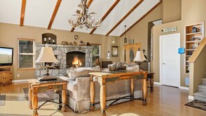 A living room with a stone fireplace, wooden furniture, a chandelier, and a staircase. - Large Retreat, Arcade, Lake Access + AC (Lake Arrowhead)
