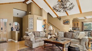 A spacious living room with high ceilings, a chandelier, and a fireplace. There is a large window providing natural light and a view of the outdoors. - Large Retreat, Arcade, Lake Access + AC (Lake Arrowhead)