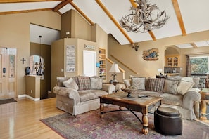 A spacious living room with high ceilings, a chandelier, and a fireplace. There is a large window providing natural light and a view of the outdoors.