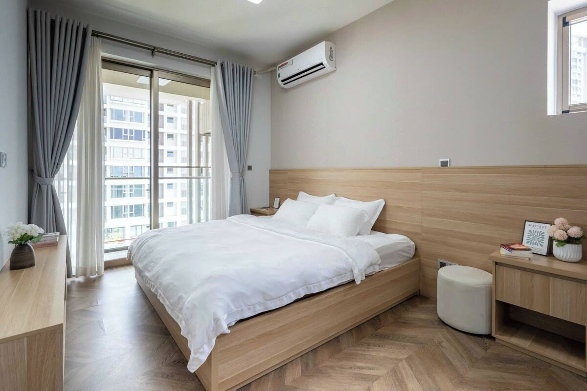 Quarto de hotel moderno com cama de madeira, mesa de cabeceira, um banquinho branco pequeno e janela com cortinas.