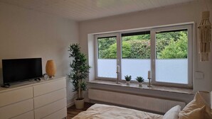 Un dormitorio con cama, televisor en una cómoda, ventana con persianas y una planta en maceta.