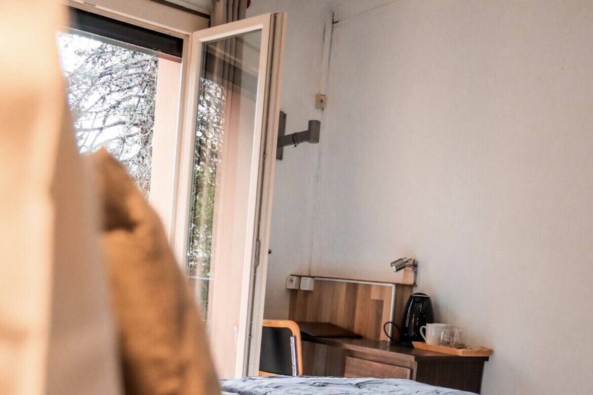 A bedroom with a bed, a wooden desk, a coffee maker, and a window with a view of trees.