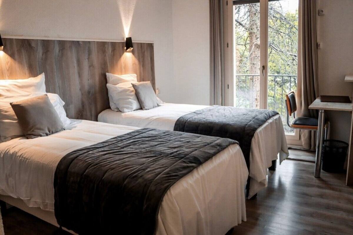 A bedroom with two beds, a wooden headboard, a desk, and a view of trees through the window.