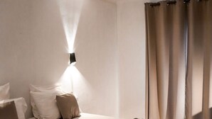 Una cama bien hecha con un edredón oscuro, una lámpara de pared y una ventana con cortinas.