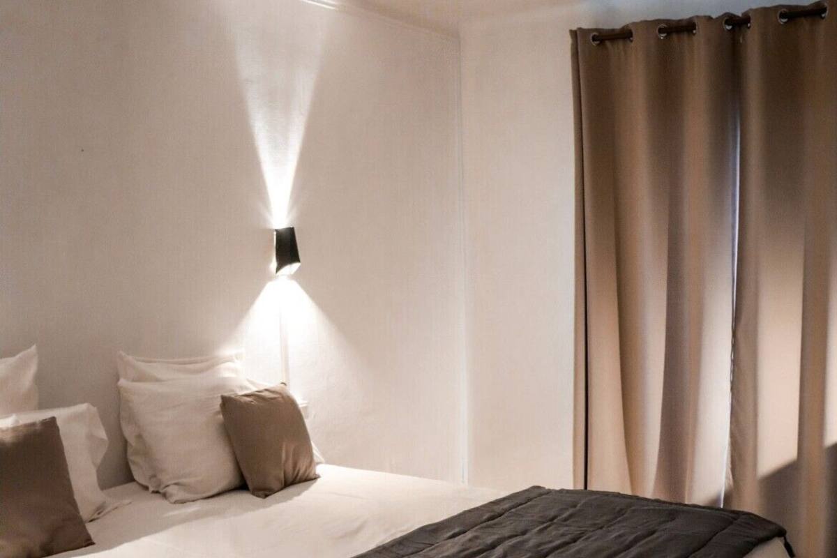 A neatly made bed with a dark blanket, a wall-mounted light, and a window with curtains.