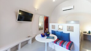 A modern, compact room with a white bench, a round table with a bowl and book, a sofa with colorful cushions, and a mounted TV.