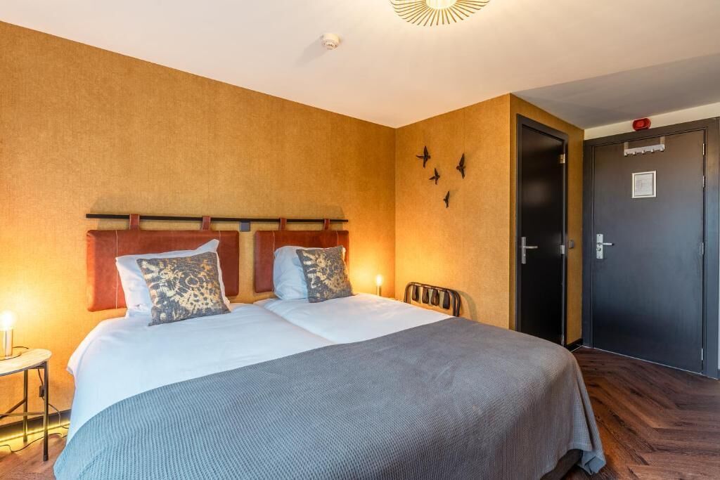 Una cama doble con cabecera de madera, dos mesitas de noche con lámparas, una puerta oscura y un aplique en la pared.