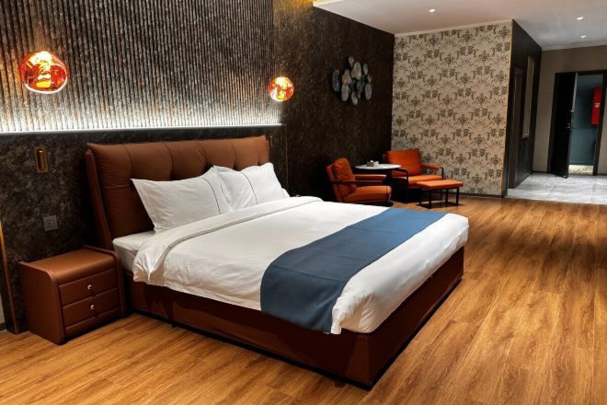A modern hotel room with a large bed, a nightstand, a seating area with a table and chairs, and a decorative wall.