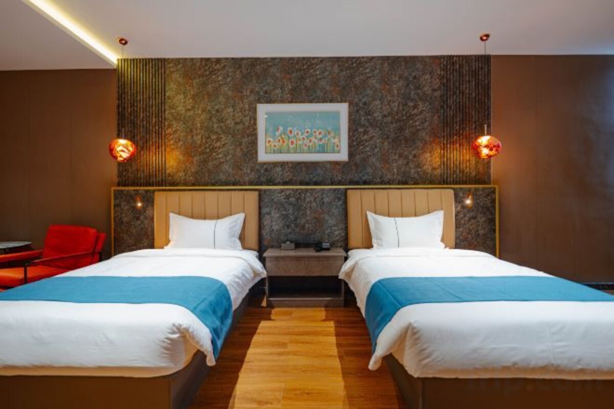 A hotel room with two beds, a wooden floor, a painting on the wall, and a red chair.