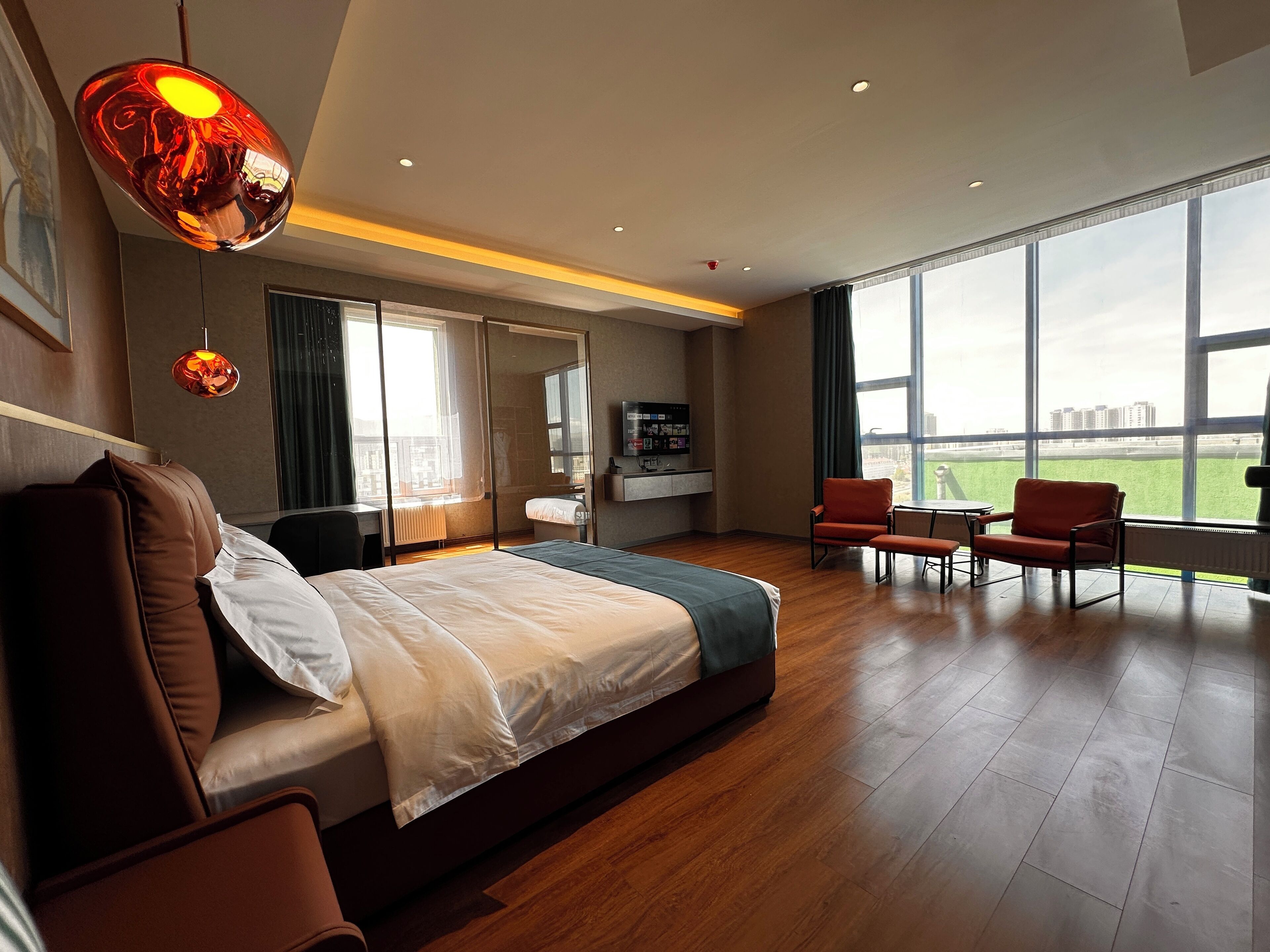 A modern hotel room with a large bed, a desk, a chair, and a TV. The room has large windows offering a view of the city.