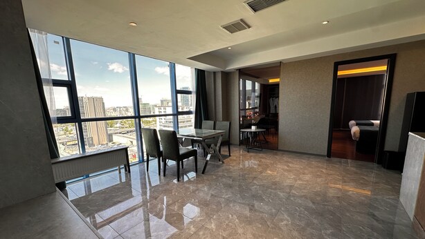 Una habitación de hotel moderna con un ventanal grande que ofrece vistas a la ciudad, una zona de comedor con mesa y sillas, y una zona de descanso separada con cama.