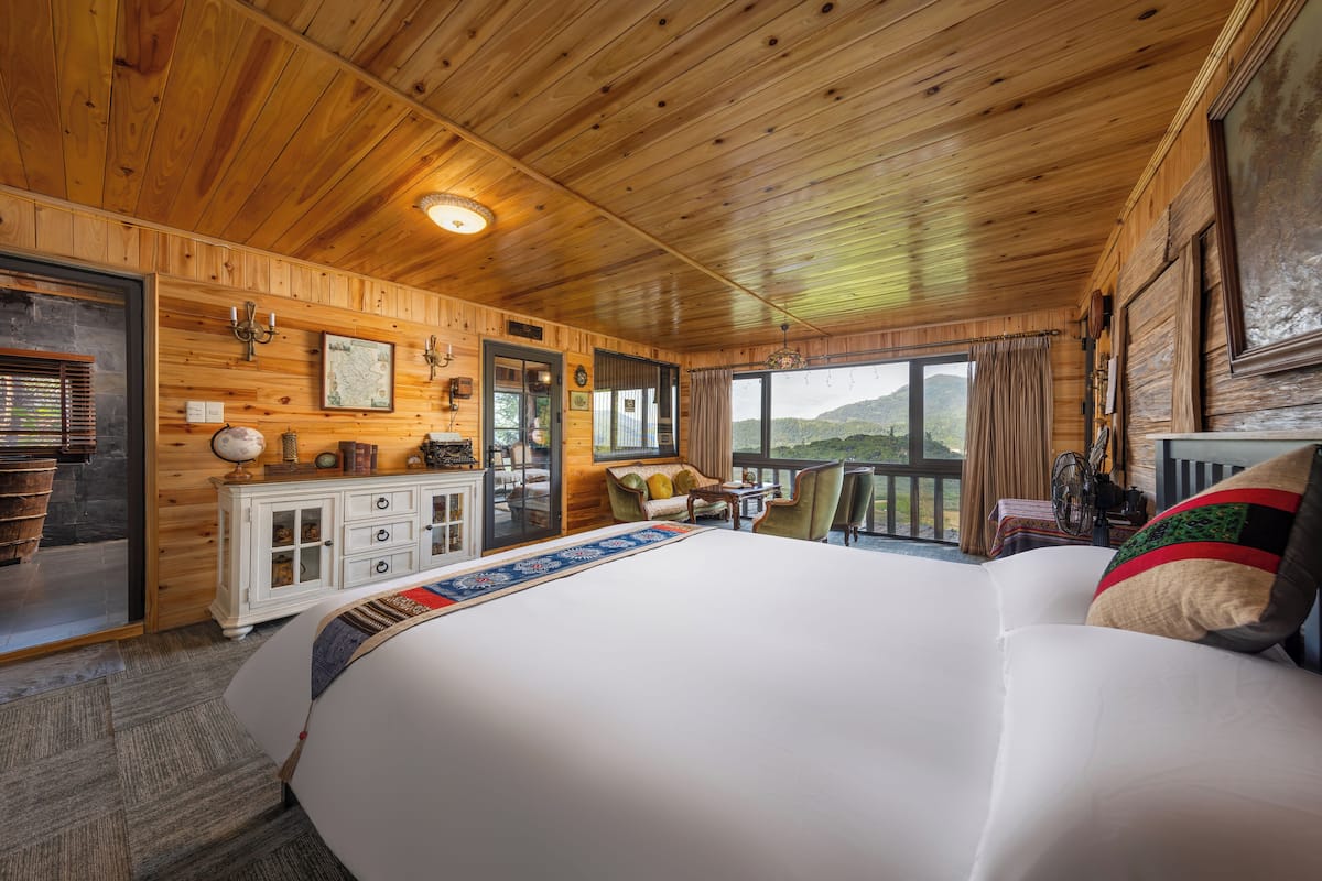 A wooden cabin room with a large bed, a wooden dresser, a dining table with chairs, and a view of the outdoors.