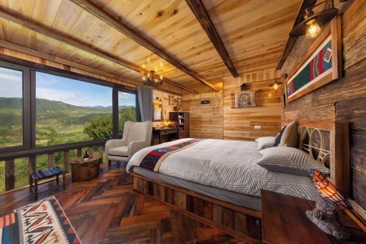 A wooden cabin with a large bed, a desk, and a view of the landscape.
