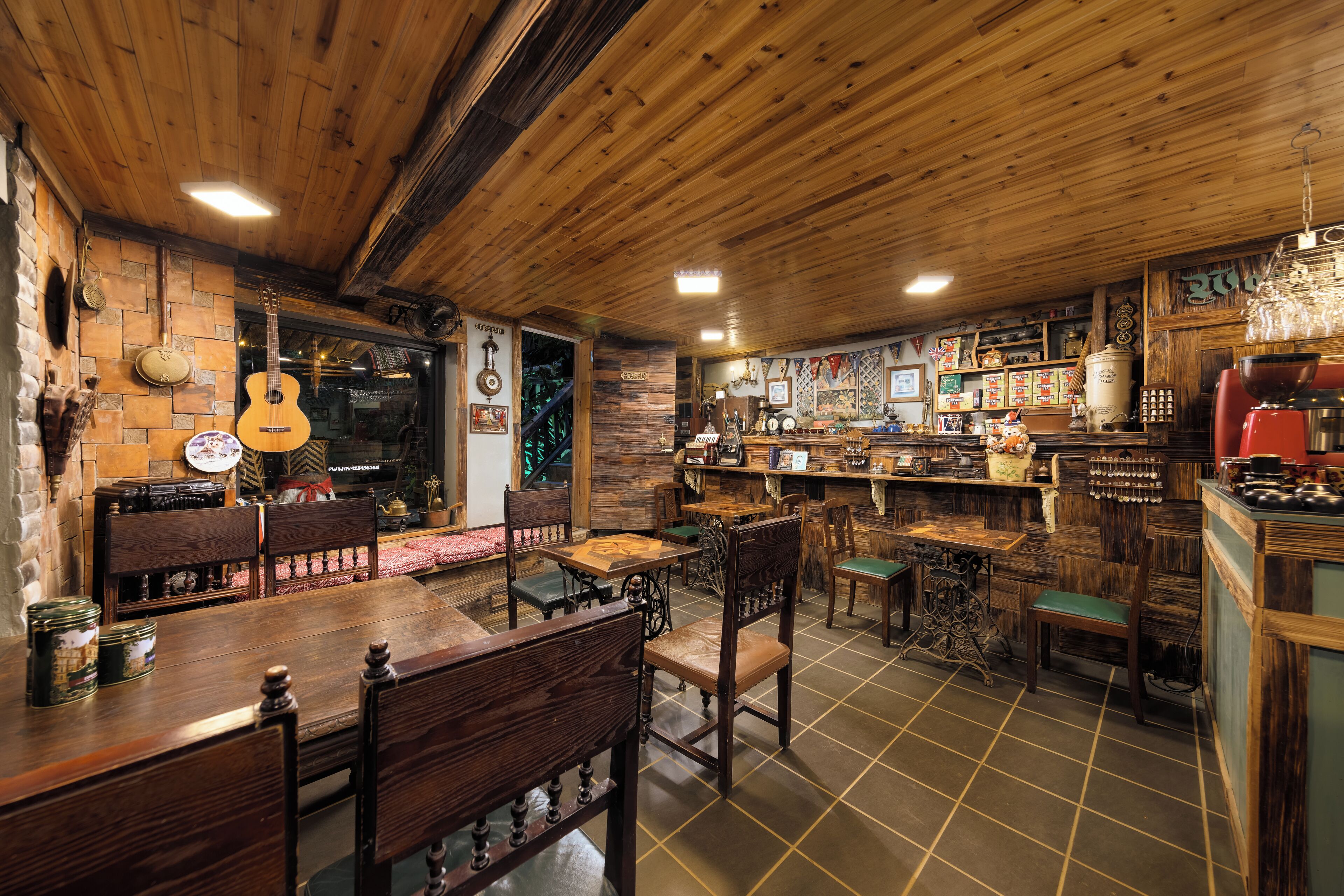 A rustic interior with wooden walls and ceiling, a bar area with stools and a counter, and a dining area with wooden tables and chairs.