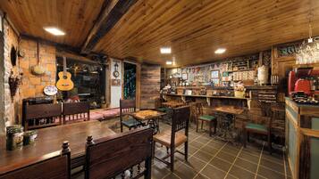 A rustic interior with wooden walls and ceiling, a bar area with stools and a counter, and a dining area with wooden tables and chairs.