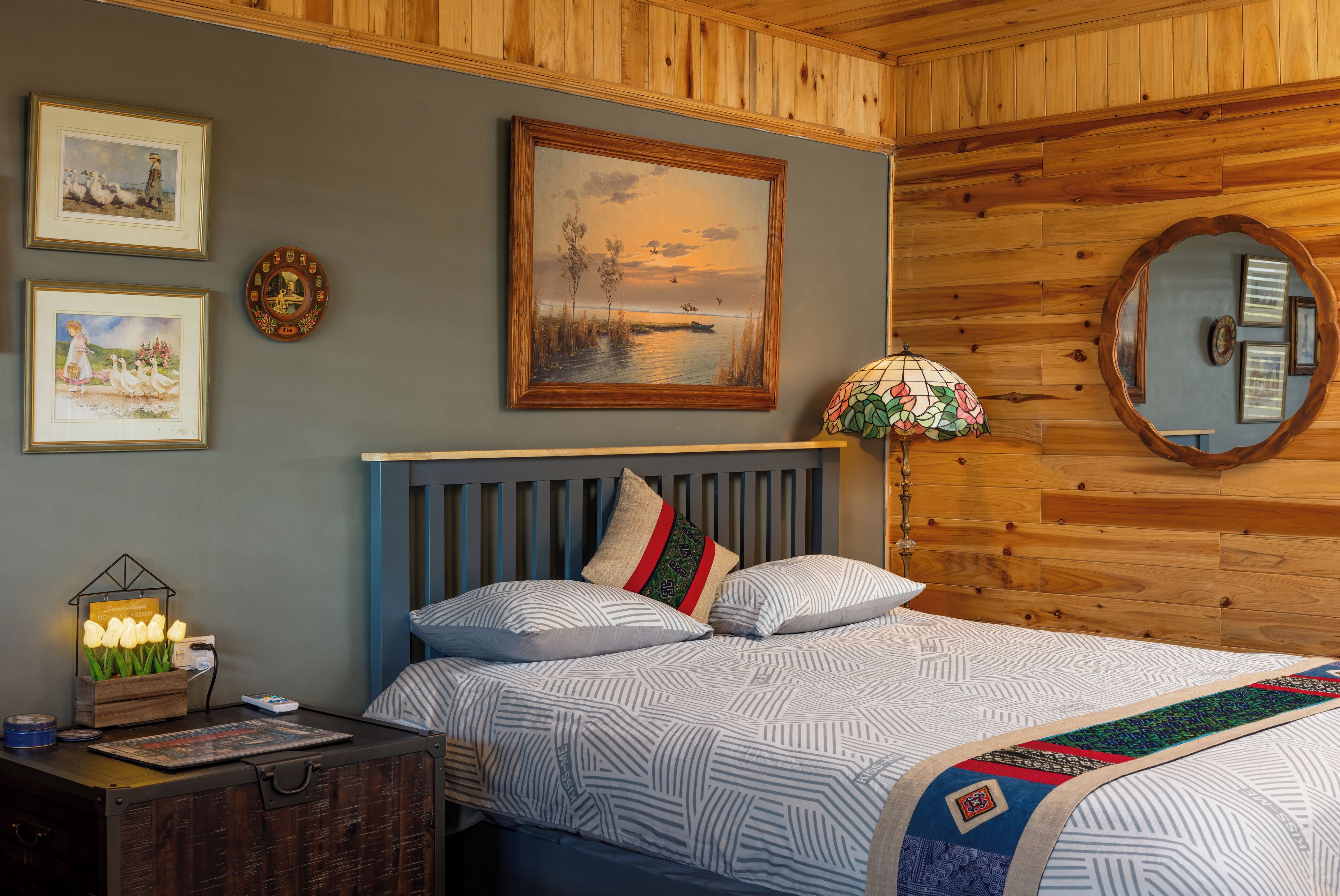 A bedroom with a wooden headboard, a bed with patterned bedding, a nightstand with a lamp, and wall art including a framed painting and a mirror.