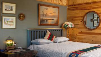 A bedroom with a wooden headboard, a bed with patterned bedding, a nightstand with a lamp, and wall art including a framed painting and a mirror.