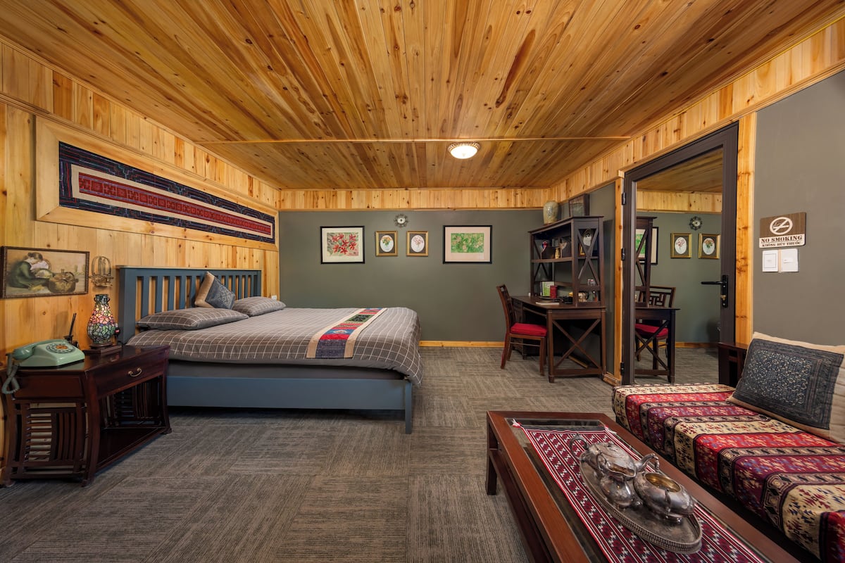 A room with a wooden ceiling, a bed with a blue headboard, a desk with a chair, and a sofa with patterned cushions.