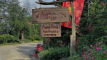 A wooden sign for 'Captain's Cottage' with a food and drink service, surrounded by lush greenery and a red flag.
