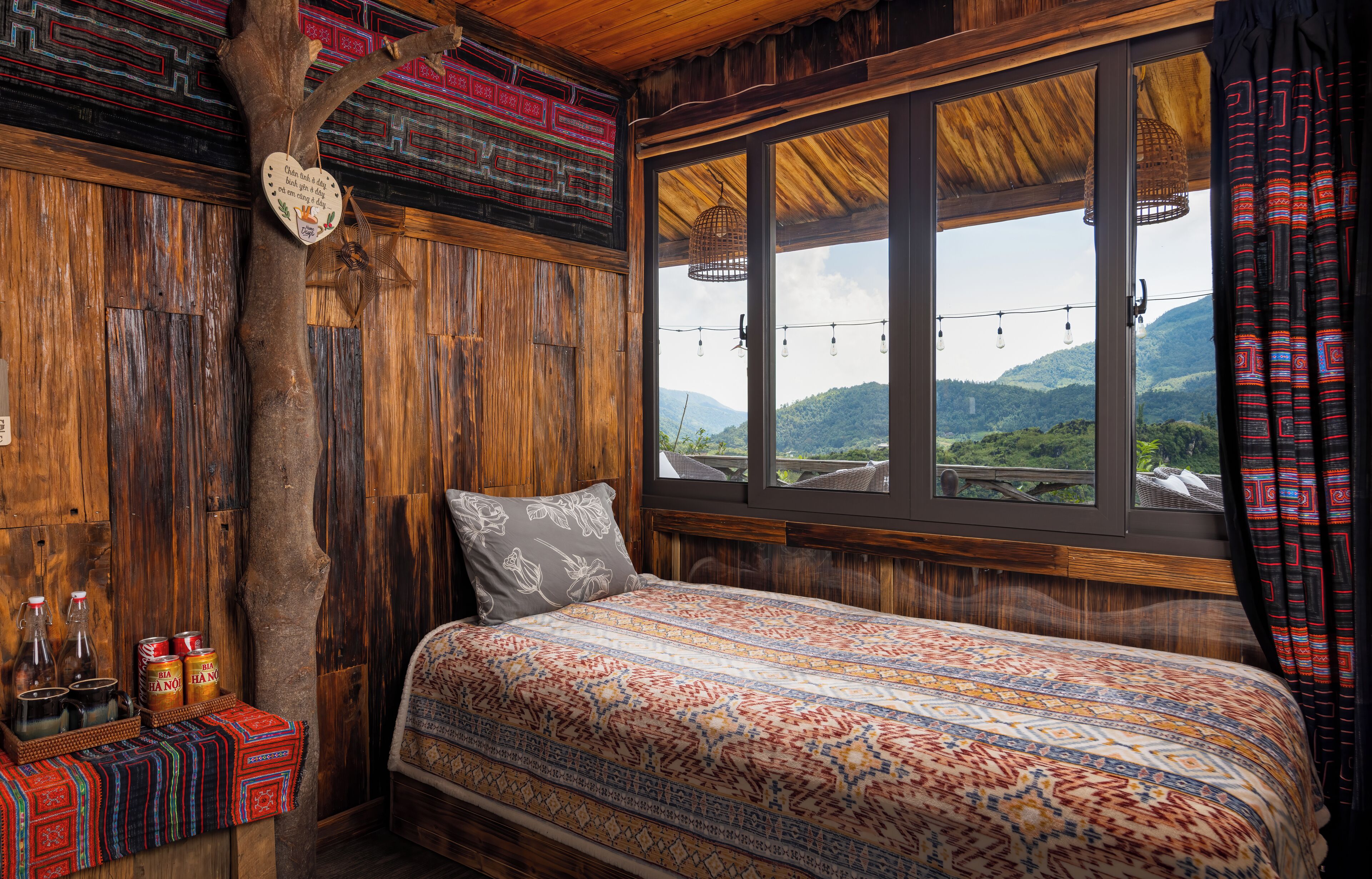 A bedroom with a large window, a bed with patterned bedding, wooden walls, and a tree branch decoration.